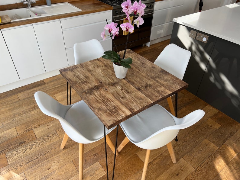 May include: A square wooden dining table with black hairpin legs and four white chairs. A potted orchid with pink flowers sits in the center of the table.