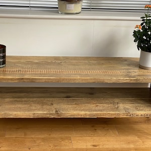 Puede incluir: Una mesa de centro de madera r&uacute;stica con un estante inferior y patas de horquilla negras. La mesa tiene un acabado de madera natural y est&aacute; decorada con una vela encendida y una planta en maceta con flores naranjas.