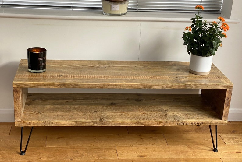 Puede incluir: Una mesa de centro de madera r&uacute;stica con un estante inferior y patas de horquilla negras. La mesa tiene un acabado de madera natural y est&aacute; decorada con una vela encendida y una planta en maceta con flores naranjas.
