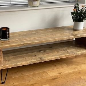 May include: A rustic wooden coffee table with a lower shelf and black hairpin legs. The table has a natural wood finish and is made from reclaimed wood. The table is 120 cm long and 40 cm wide.