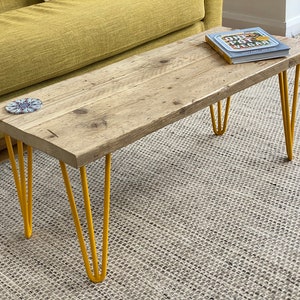 May include: A rustic wooden coffee table with a natural wood finish and yellow hairpin legs. The table is sitting on a light grey rug. A cookbook titled "One Pot Vegan" is on top of the table.