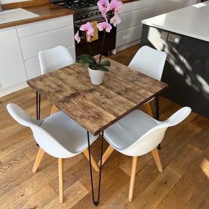 May include: A square wooden dining table with black hairpin legs and four white chairs. The table has a rustic wood grain finish. A potted orchid plant sits in the center of the table.