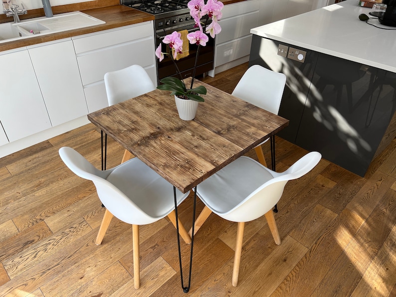 May include: A square wooden dining table with black hairpin legs and four white chairs. The table has a rustic wood grain finish. A potted orchid plant sits in the center of the table.