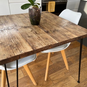 May include: A square wooden table with a rustic finish and black hairpin legs. The table is set with two white chairs and a potted plant.
