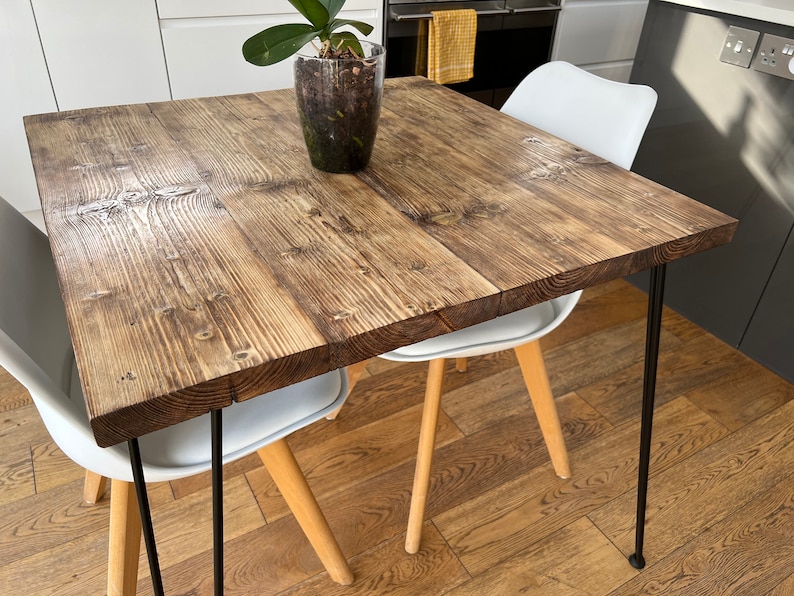 May include: A square wooden table with a rustic finish and black hairpin legs. The table is set with two white chairs and a potted plant.