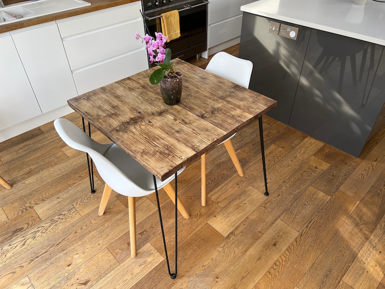 May include: A square wooden table with black hairpin legs and a light wood finish. The table is set with a white chair and a potted orchid with pink flowers.