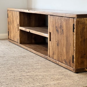 May include: A low, wooden media unit with a natural finish. It has two sets of doors and an open shelf. The wood grain is visible, enhancing its rustic charm. The unit is designed to sit close to the floor, with a length of approximately 180 cm.