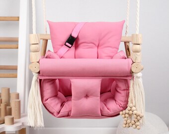 May include: Pink baby swing with a padded seat and a safety harness. The swing has wooden beads and a star-shaped wooden toy hanging from the bottom.