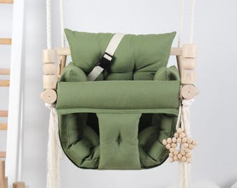 May include: A green fabric baby swing with a wooden frame and white ropes. The swing has a safety harness and a wooden star toy.