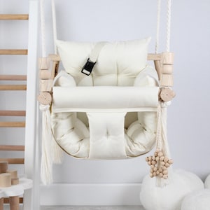 May include: A white padded swing seat with a wooden frame and rope straps. The swing has a safety belt and a wooden star-shaped toy hanging from the bottom.