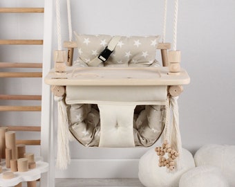 May include: A wooden baby swing with a beige padded seat and a tray. The swing has white ropes and a white star-shaped bead toy hanging from the bottom. The swing is in a room with a wooden ladder and white fluffy pillows.