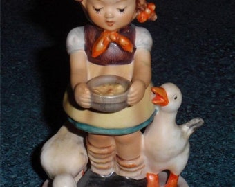 May include: A porcelain figurine of a girl in a brown dress and orange scarf feeding a white goose from a bowl. The girl is standing on a white base with two white geese.