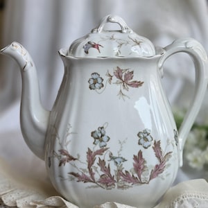 May include: A white porcelain teapot with a fluted body and a matching lid. The teapot features a delicate botanical pattern of light blue flowers and muted pinkish-brown leaves. It has a curved handle, a shaped spout, and gold trim accents.