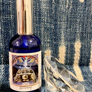 Puede incluir: Una botella de vidrio azul con una tapa plateada y una etiqueta que dice "Grounding". La etiqueta presenta una ilustración colorida de una tortuga con una luna creciente y un pájaro con las alas extendidas. Un diamante de cristal transparente está al lado de la botella.