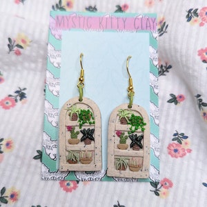 Pode incluir: Um par de brincos pendentes com uma janela de cerâmica branca com um design de plantas verdes e marrons. Os brincos estão em um cartão que diz "Mystic Kitty Clay".