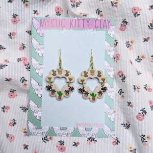 May include: A pair of heart-shaped earrings made of clay with a green and brown succulent plant design. The earrings are hanging from gold hooks. The earrings are on a white background with a pink and green pattern.