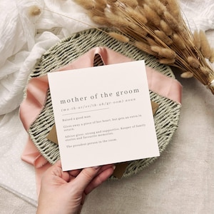 Puede incluir: Una tarjeta cuadrada con el texto "mother of the groom" se muestra en una cesta tejida con una cinta rosa. La tarjeta incluye una definición y descripción del rol. Flores secas en el fondo.