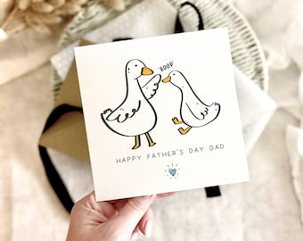 May include: A greeting card with a cartoon illustration of two white ducks, one saying "boop" to the other. The text "HAPPY FATHER'S DAY DAD" is printed below the ducks, with a blue heart. The card is held by a hand.