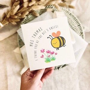 May include: A square white greeting card featuring a cartoon bee with a heart-shaped wing, pink flowers, and the black printed text: MRS THOMAS I THINK YOU'RE THE BEE'S KNEES! The design uses simple lines and soft colours on a textured paper surface.