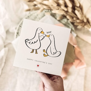 K&ouml;nnte beinhalten: Eine quadratische Valentinstagskarte mit zwei Cartoon-Enten, von denen eine 'boop' sagt. Die Karte tr&auml;gt den Text 'Happy Valentine's Day' und ein kleines rotes Herz. Die Karte wird von einer Hand gehalten.