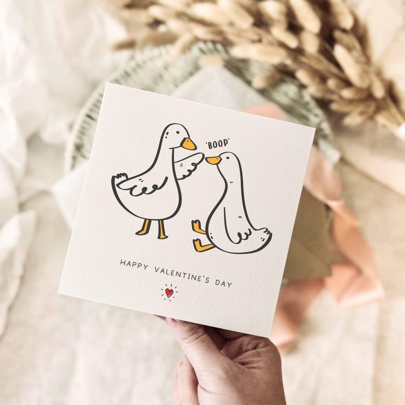 K&ouml;nnte beinhalten: Eine quadratische Valentinstagskarte mit zwei Cartoon-Enten, von denen eine 'boop' sagt. Die Karte tr&auml;gt den Text 'Happy Valentine's Day' und ein kleines rotes Herz. Die Karte wird von einer Hand gehalten.