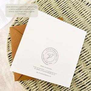 A white greeting card with a brown envelope, on a woven surface. The card has a circular logo with a bird design and the text "LITTLE PAPER BIRD LTD". Text on the card states the cards are printed on a thick textured card stock.