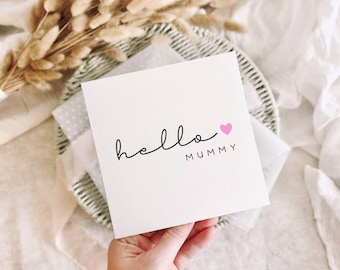 May include: A white card with the text "hello MUMMY" in black script and a pink heart. The card is held in a hand and is placed on a woven plate. Dried flowers are in the background.