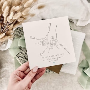 May include: A white Father's Day card with a line drawing of a hand holding three smaller hands. The card reads "HAPPY FATHER'S DAY we love you EST. 2015" with the names "Jude", "Evan", and "Adeline" written on the card.