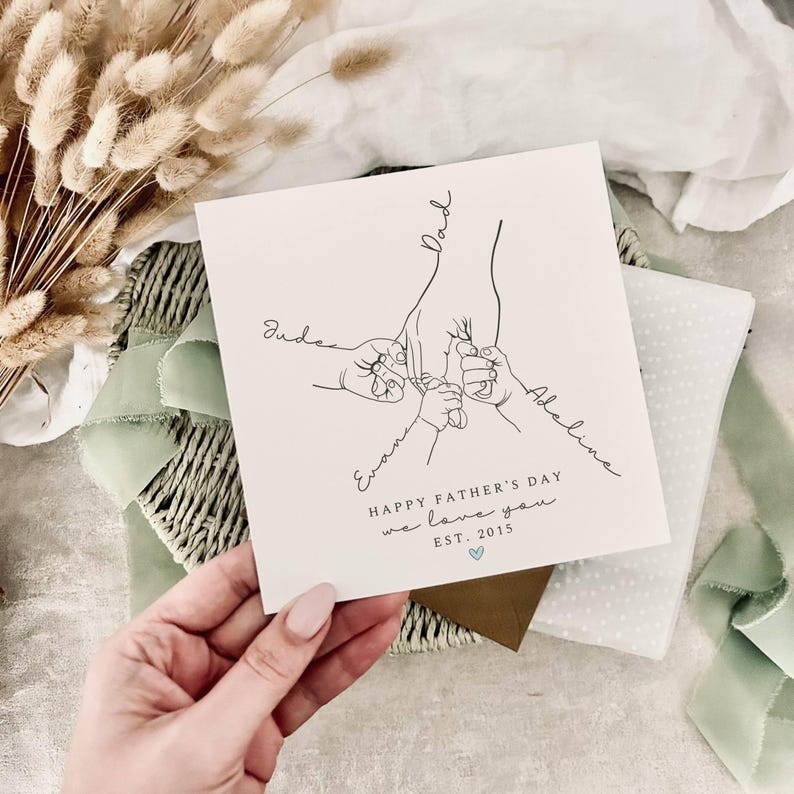 May include: A white Father's Day card with a line drawing of a hand holding three smaller hands. The card reads "HAPPY FATHER'S DAY we love you EST. 2015" with the names "Jude", "Evan", and "Adeline" written on the card.