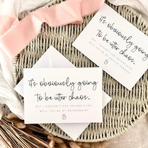 Może przedstawiać: Dwie białe karty z czarnym napisem "It's obviously going to be utter chaos..." są wyświetlane na tkanej tacy. Na kartach widnieją pytania "Will you be my bridesmaid?" i "Will you be my maid of honour?" Różowa wstążka jest ułożona na tacy.