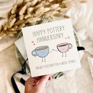 May include: A square white greeting card featuring the text HAPPY POTTERY ANNIVERSARY and BREWED TOGETHER FOR 9 WHOLE YEARS. The design includes a light blue mug and a light pink mug with smiling faces, stick arms, and legs holding hands.