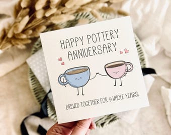 May include: A square white greeting card featuring the text HAPPY POTTERY ANNIVERSARY and BREWED TOGETHER FOR 9 WHOLE YEARS. The design includes a light blue mug and a light pink mug with smiling faces, stick arms, and legs holding hands.