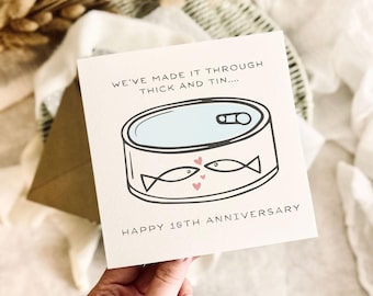 Könnte beinhalten: Eine quadratische Grußkarte mit weißem Hintergrund und einer Cartoon-Illustration einer Konservendose mit zwei Fischen, die sich gegenüberstehen, sowie kleinen rosa Herzen. Der Text lautet: WE'VE MADE IT THROUGH THICK AND TIN.... HAPPY 10TH ANNIVERSARY.