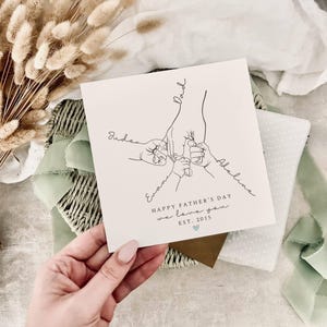 A white greeting card with the text "Happy Father's Day, We Love You" printed on the bottom centre of the card.  The image above is line drawn hands (1 adult and 3 children). The children's hands are holding the adult fingers.  The card is personalised with the children's names connected to each hand.