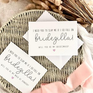 Puede incluir: Dos tarjetas blancas con texto en negro que dice: "I NEED YOU TO SLAP ME IF I GO FULL ON bridezilla! WILL YOU BE MY BRIDESMAID?" y "I NEED YOU TO SLAP ME IF I GO FULL ON bridezilla! WILL YOU BE MY MAID OF HONOUR?" Un corazón rosa está debajo del texto. Las tarjetas están en una cesta tejida.