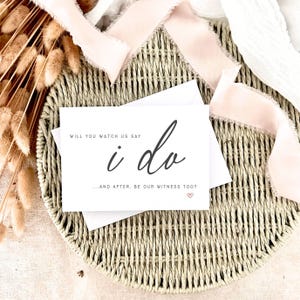 Puede incluir: Una tarjeta de invitación blanca con las palabras "i do" en escritura elegante, sobre una cesta tejida. La tarjeta también incluye el texto "Will you watch us say" y "...and after, be our witness too?" con un pequeño corazón.
