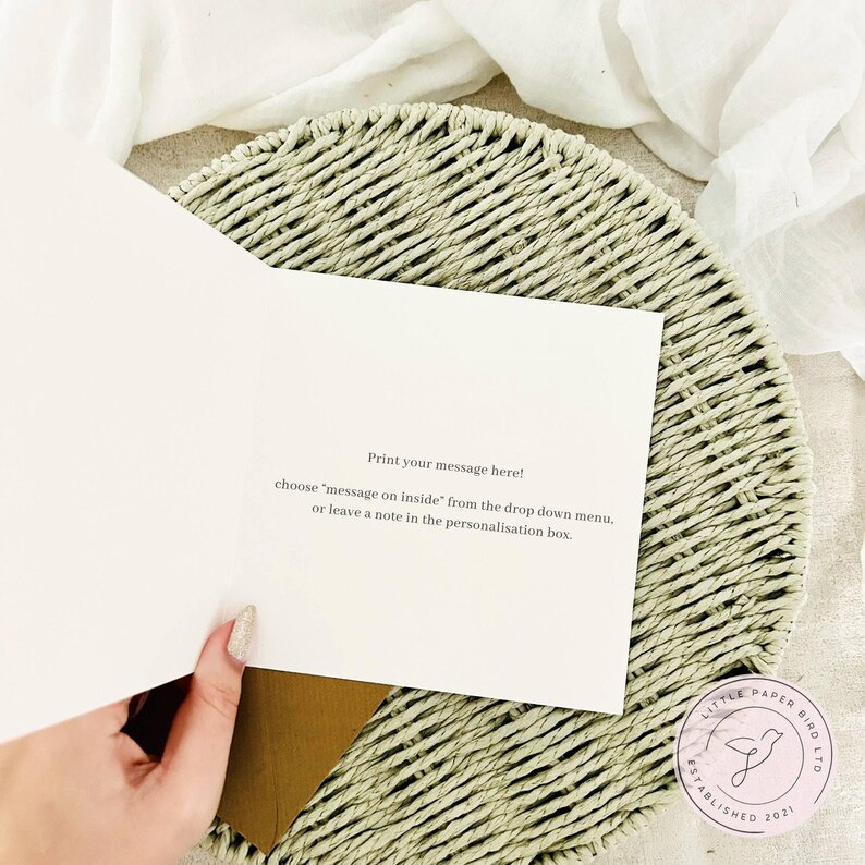 K&ouml;nnte beinhalten: Eine offene Gru&szlig;karte mit dem Text "Print your message here!". Die Karte wird &uuml;ber einem geflochtenen Platzset und einem braunen Umschlag gehalten. Das Logo von Little Paper Bird Ltd befindet sich in der unteren rechten Ecke. Das Innere der Karte ist leer.
