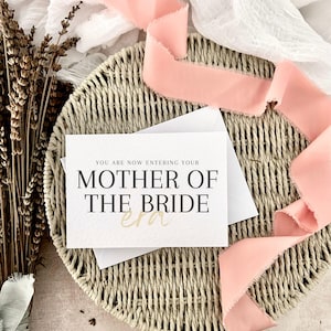 May include: A white rectangular greeting card with black serif text that reads YOU ARE NOW ENTERING YOUR MOTHER OF THE BRIDE and the word era in a gold script font. The card is placed on a round woven wicker tray with a long peach silk ribbon.