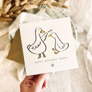 May include: A square white greeting card featuring a black and white line drawing of two ducks. One duck is touching the other on the beak with the text BOOP above them. Below the ducks, the text reads HAPPY BIRTHDAY DADDY with a small blue heart.