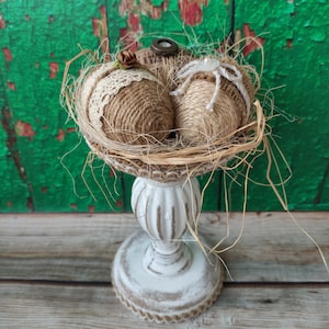 Może przedstawiać: Dekoracyjny element centralny z dwiema kulami owiniętymi sznurkiem w gnieździe z plecionki, umieszczony na białym, ozdobnym cokole. Ozdobiony jest suchą trawą, małym kwiatkiem i guzikiem. Podstawa ma średnicę około 15 cm.