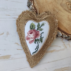 Op de afbeelding: Een hartvormig hangend ornament met een centraal wit stoffen paneel met een in kruissteek geborduurde roze roos en groene bladeren. Het hart is omrand met gerafelde bruine jute stof en bevat een klein strikje van touw aan de bovenkant.