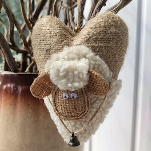 Peut inclure: Ornement en forme de cœur avec un motif de mouton. Le cœur est enveloppé de ficelle, avec un visage de mouton blanc duveteux et des oreilles en toile de jute. Une petite cloche est suspendue en dessous. Décoration artisanale.
