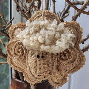 May include: A handmade burlap sheep ornament with a white fluffy wool head and black eyes. The sheep has a brown burlap body with a curly tail and is hanging from a string.