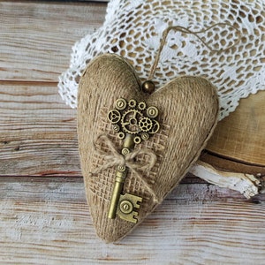 Peut inclure: Ornement en forme de cœur enveloppé de ficelle, avec un centre en toile de jute. Une clé en bronze avec des accents d'engrenages est fixée à l'avant. Une petite boucle et une perle sont attachées pour suspendre. L'ornement repose sur une surface en bois.
