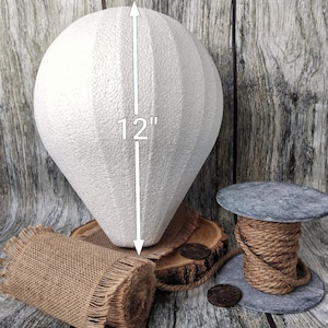 May include: A white, teardrop-shaped foam craft form, approximately 12 inches tall, with vertical textured lines. The form rests on a wooden slice and burlap. A spool of rope and a decorative wooden disc are also present.