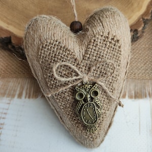 Peut inclure: Ornement en forme de cœur en toile de jute avec un nœud en jute et un charm en forme de hibou en bronze. L'ornement est suspendu par une ficelle en jute et une perle en bois. L'arrière-plan comprend une tranche de bois et plus de toile de jute.