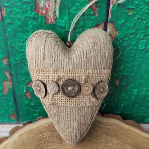 Peut inclure: Ornement en forme de cœur fait de ficelle, avec une bande de toile de jute ornée de boutons en bois. La ficelle est enroulée autour du cœur, créant une surface texturée. Une ficelle de jute est attachée pour suspendre. L'ornement est présenté sur une tranche de bois.