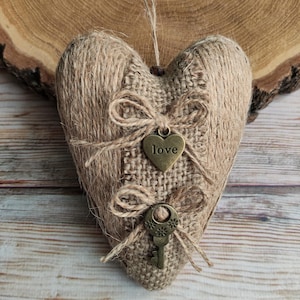 May include: A heart-shaped burlap ornament with twine detailing. It features a bronze heart charm with the word "love" and a bronze key charm. The ornament is made from woven burlap and twine, giving it a natural, rustic appearance.