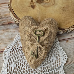 Può includere: Un ornamento a forma di cuore avvolto in spago di juta color marrone chiaro, con un inserto quadrato centrale in tela di juta. Una chiave antica in metallo color ottone è legata al centro dell'inserto in juta con un sottile spago abbinato.