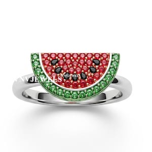 May include: A silver ring featuring a watermelon slice design. The watermelon has a red center with black seed accents, a green rind, and is encrusted with small gemstones. The ring is set against a white background.